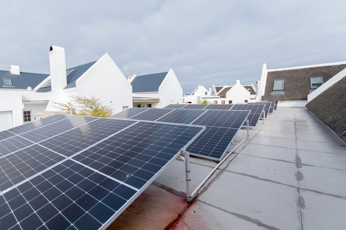 A rooftop view featuring a large array of solar panels installed on a flat roof section, with surrounding residential buildings visible.