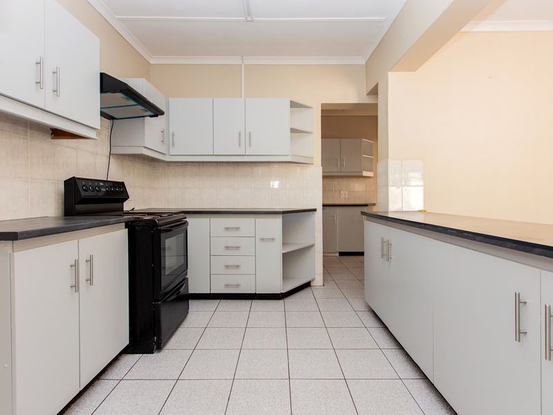 A clean, functional kitchen featuring white cabinetry, dark laminate countertops, tiled flooring, and a tiled backsplash, with an adjacent scullery area visible.