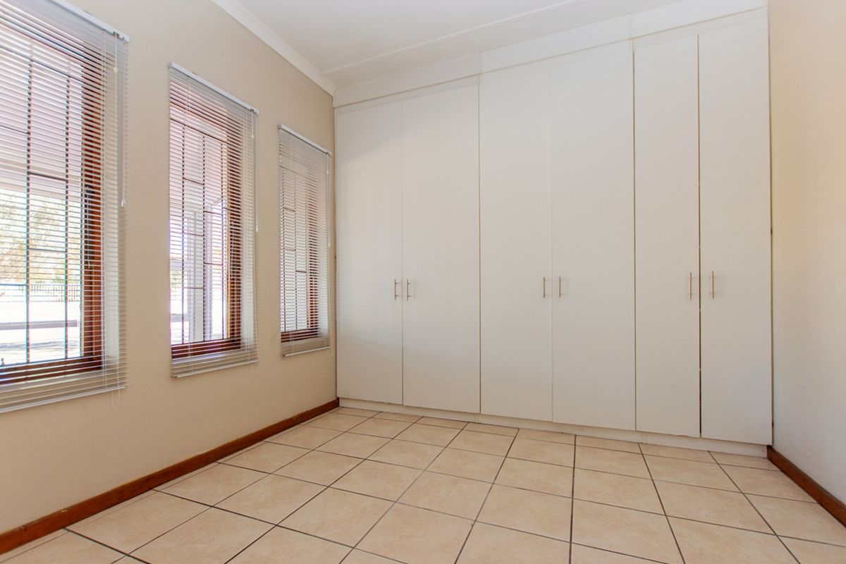 A clean, empty bedroom featuring neutral tiled flooring, ample built-in cupboard storage, and windows fitted with venetian blinds and security bars.