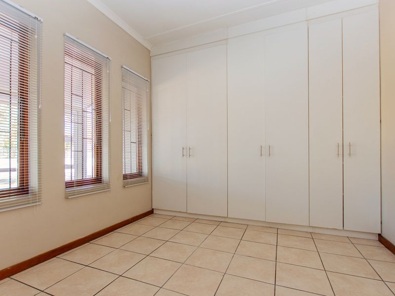 A clean, empty bedroom featuring neutral tiled flooring, ample built-in cupboard storage, and windows fitted with venetian blinds and security bars.