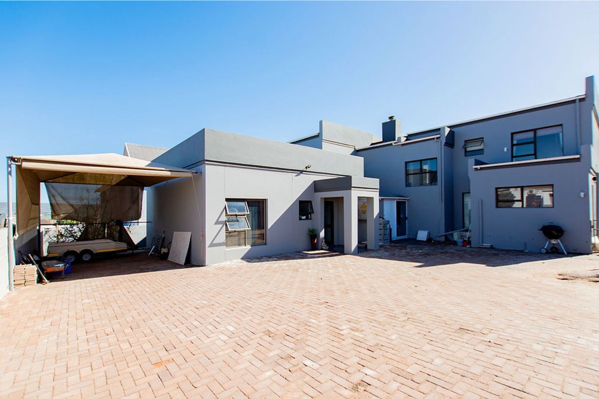 Rear exterior view of the property featuring a large paved courtyard, covered patio area, and modern grey rendered facade with aluminium window frames.