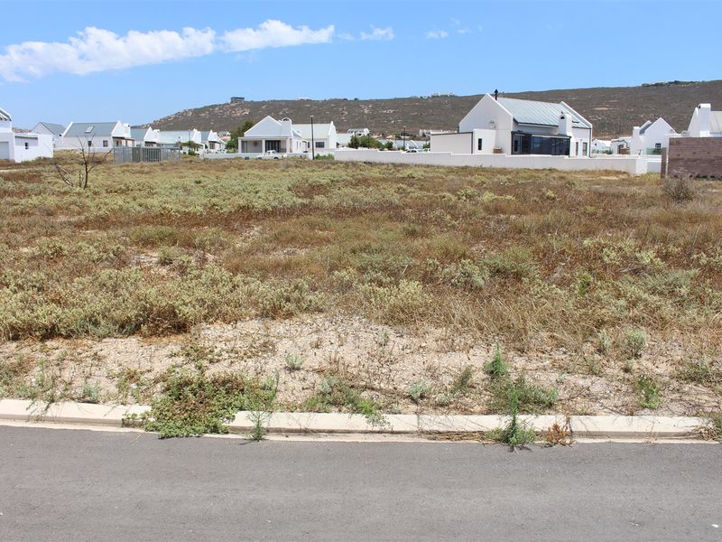 A vacant residential plot of land featuring natural vegetation, situated in a developed coastal estate with paved road access.