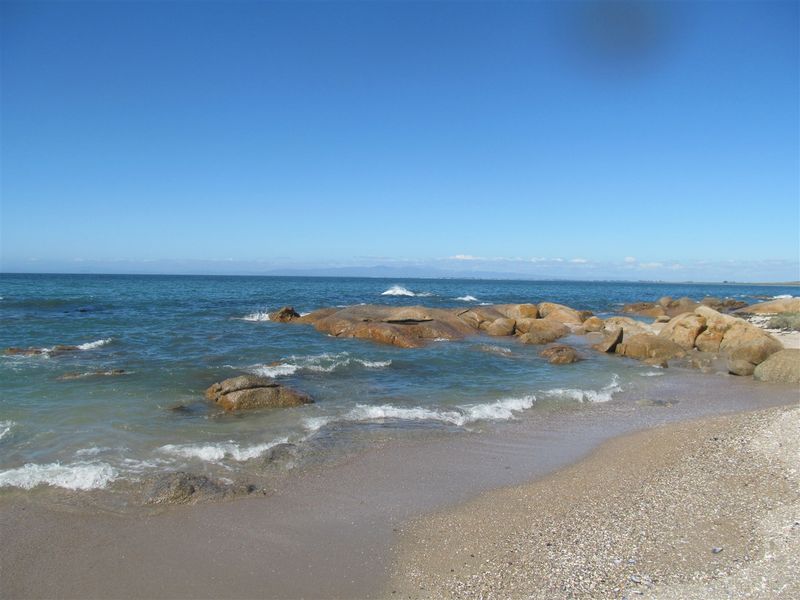 A scenic view of the coastline featuring sandy beach, rocky outcrops, and clear ocean waters under a bright blue sky.