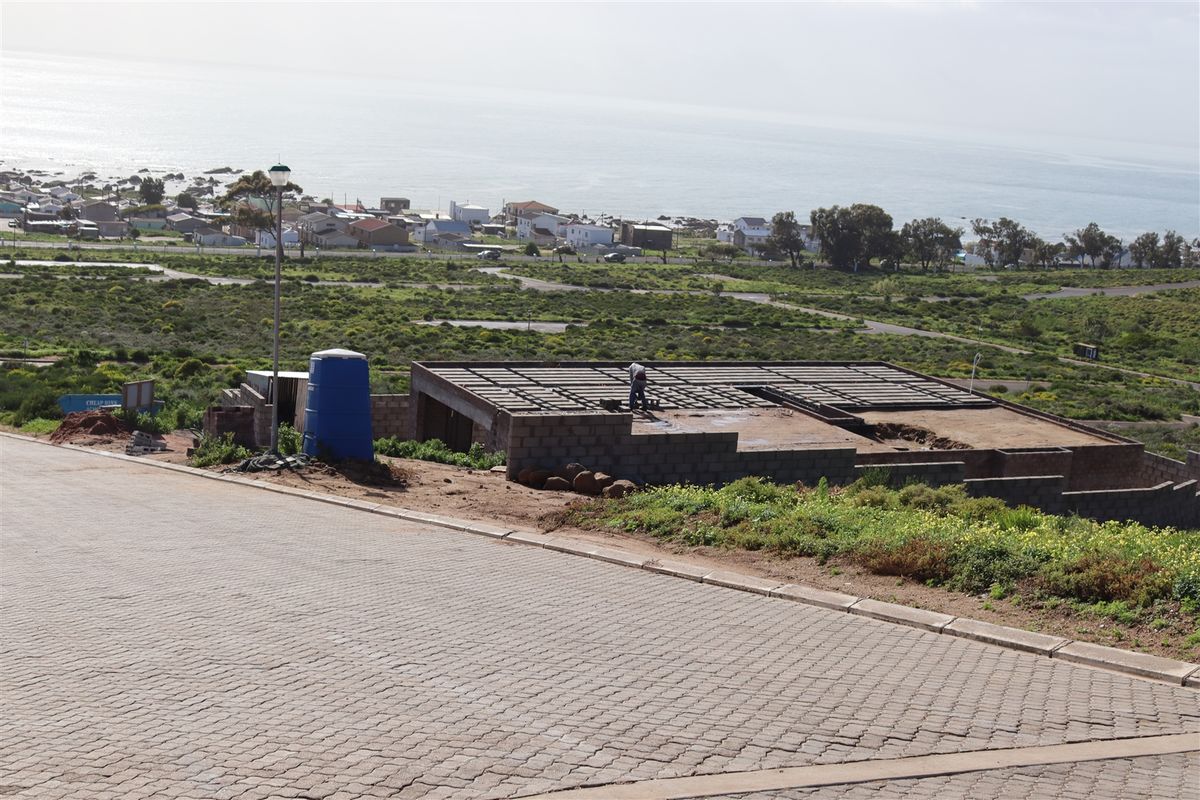 An elevated view of a residential property under construction, featuring a concrete block foundation and paved driveway with coastal views in the background.