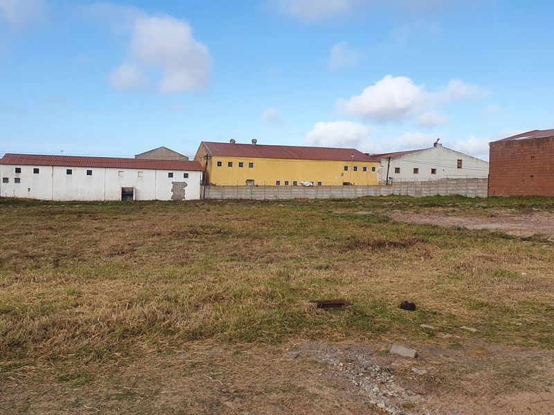 A vacant commercial land plot featuring a level grass surface, enclosed by a concrete boundary wall with adjacent industrial warehouse structures.