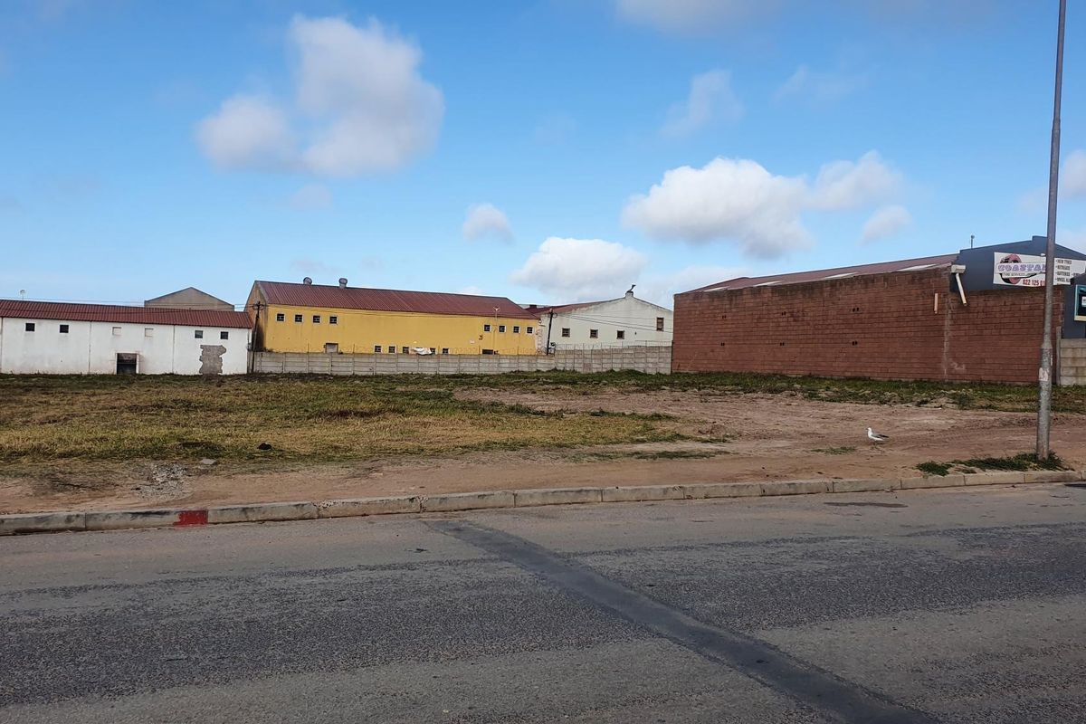 A vacant commercial land plot featuring a concrete boundary wall, situated along a paved street with surrounding industrial buildings.