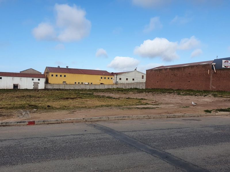 A vacant commercial land plot featuring a concrete boundary wall, situated along a paved street with surrounding industrial buildings.