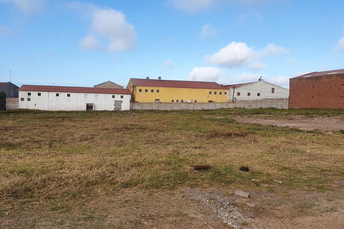 A vacant commercial land plot featuring a level grass surface, enclosed by a concrete boundary wall with adjacent industrial warehouse structures.