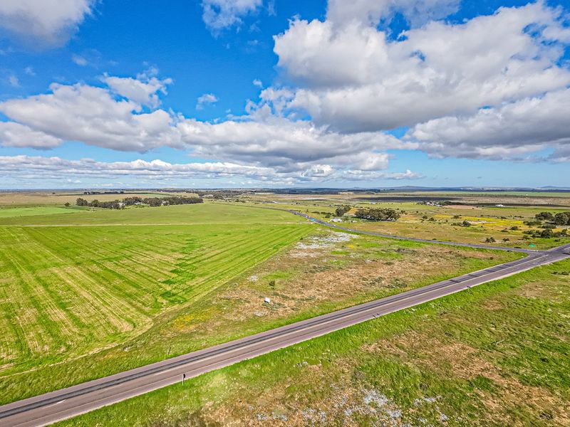 An aerial view of a large, open commercial vacant land parcel adjacent to a paved road with expansive surrounding rural landscape.