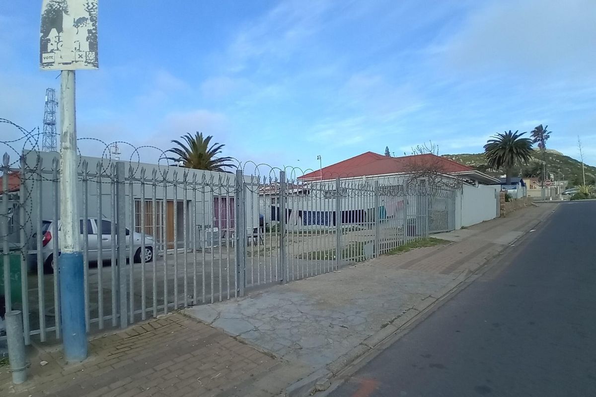 Street-level view of a commercial property featuring metal security fencing with razor wire, concrete boundary walls, and adjacent paved sidewalk.