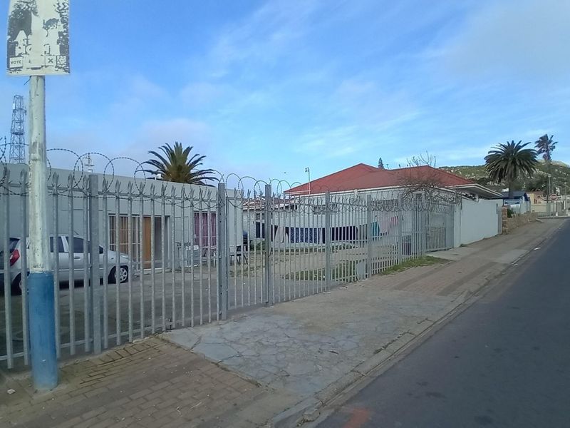 Street-level view of a commercial property featuring metal security fencing with razor wire, concrete boundary walls, and adjacent paved sidewalk.