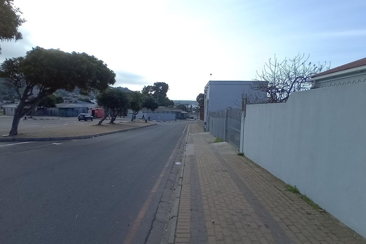 A street-level view showing the paved sidewalk, perimeter wall with security fencing, and adjacent asphalt road in a commercial area.