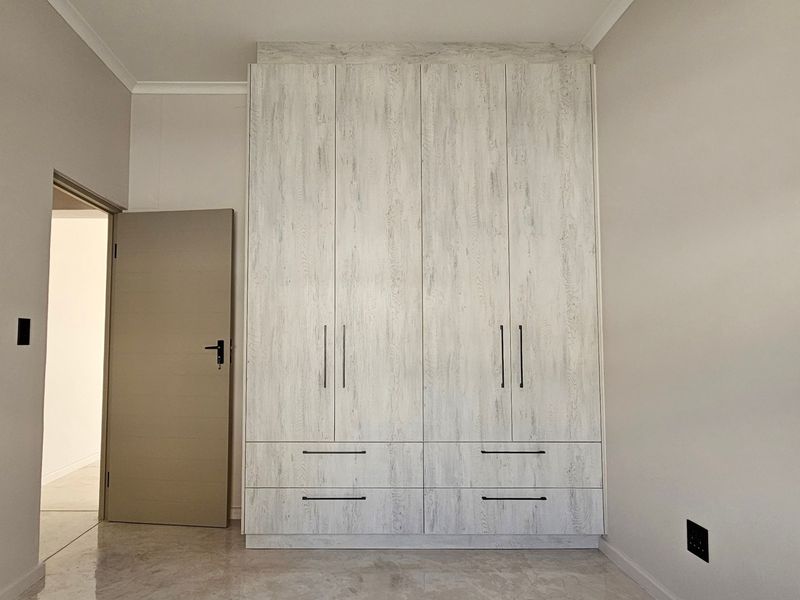 A clean, modern bedroom featuring a large, light-wood grain built-in wardrobe, neutral walls, and polished tiled flooring.
