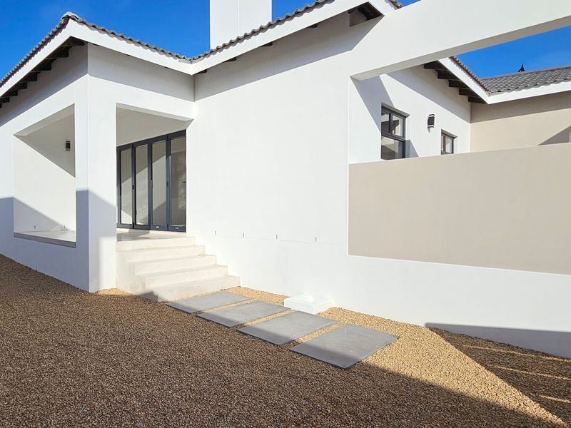 A modern rear exterior view featuring clean white rendered walls, a tiled roof, concrete steps leading to sliding glass doors, and gravel landscaping.