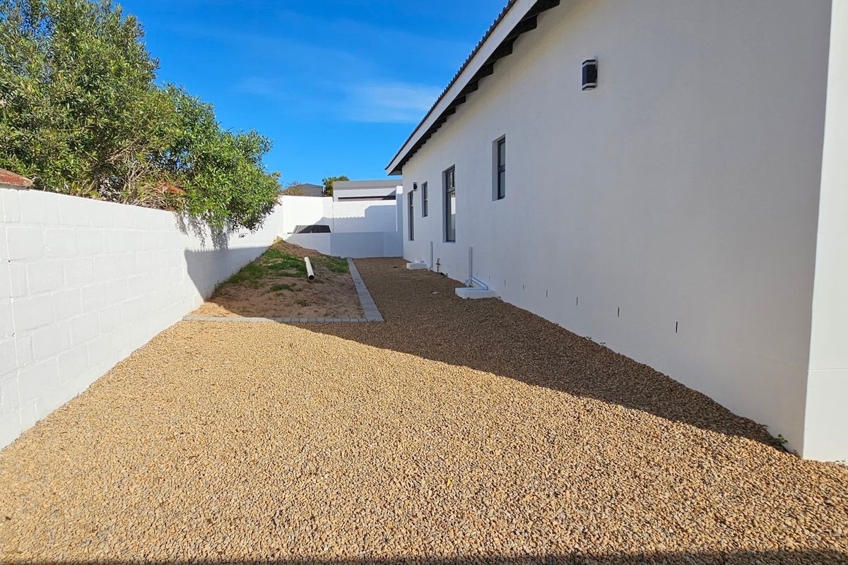 A side or rear exterior area featuring a gravel surface, boundary wall, and exterior wall lighting, with an unfinished garden section.