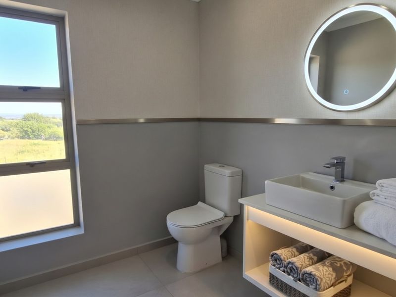 Modern bathroom featuring a floating vanity with vessel sink, back-lit circular mirror, dual-flush toilet, and large window with natural light.