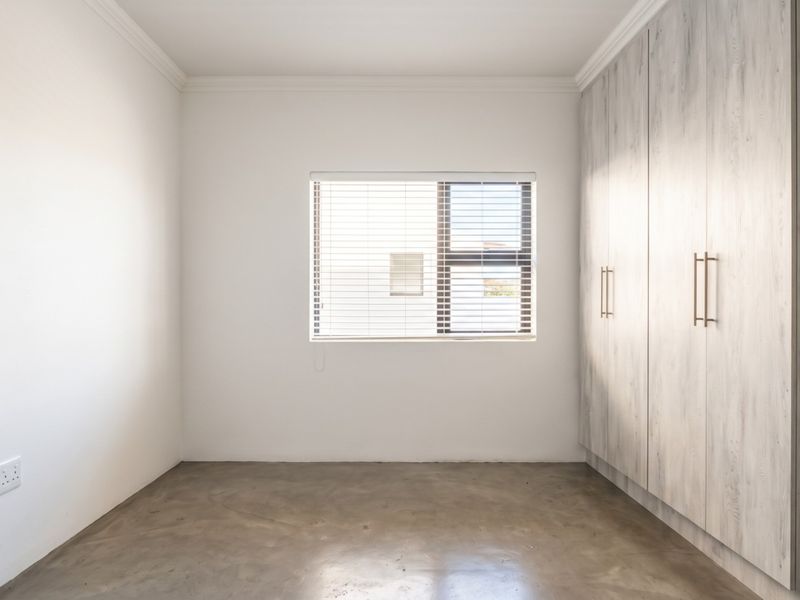 A bright, empty bedroom featuring polished concrete floors, built-in cupboards with a wood-grain finish, and venetian blinds.