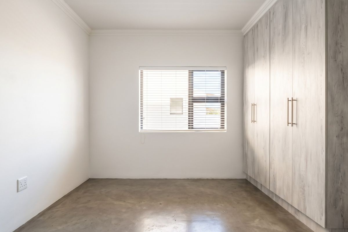 A bright, empty bedroom featuring polished concrete floors, built-in cupboards with a wood-grain finish, and venetian blinds.