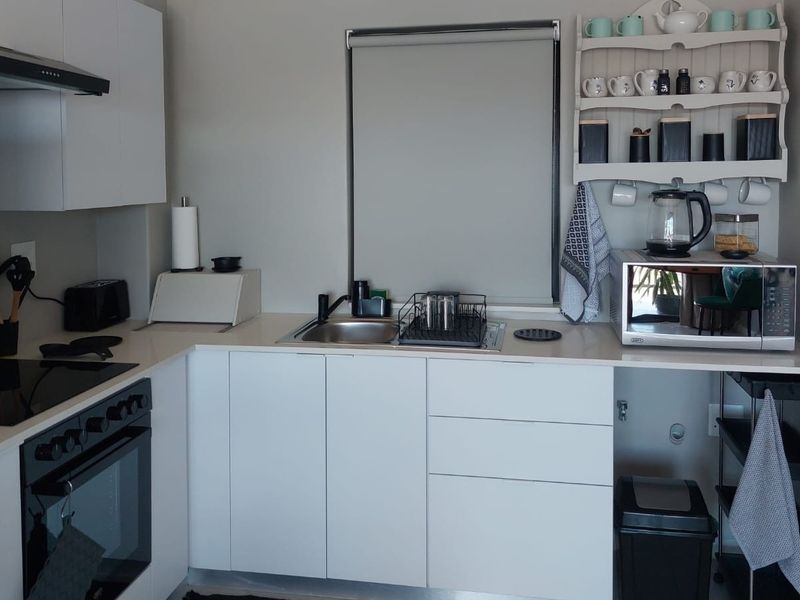 A modern, compact kitchen featuring white cabinetry, stone countertops, a built-in oven, electric hob, and a stainless steel sink.