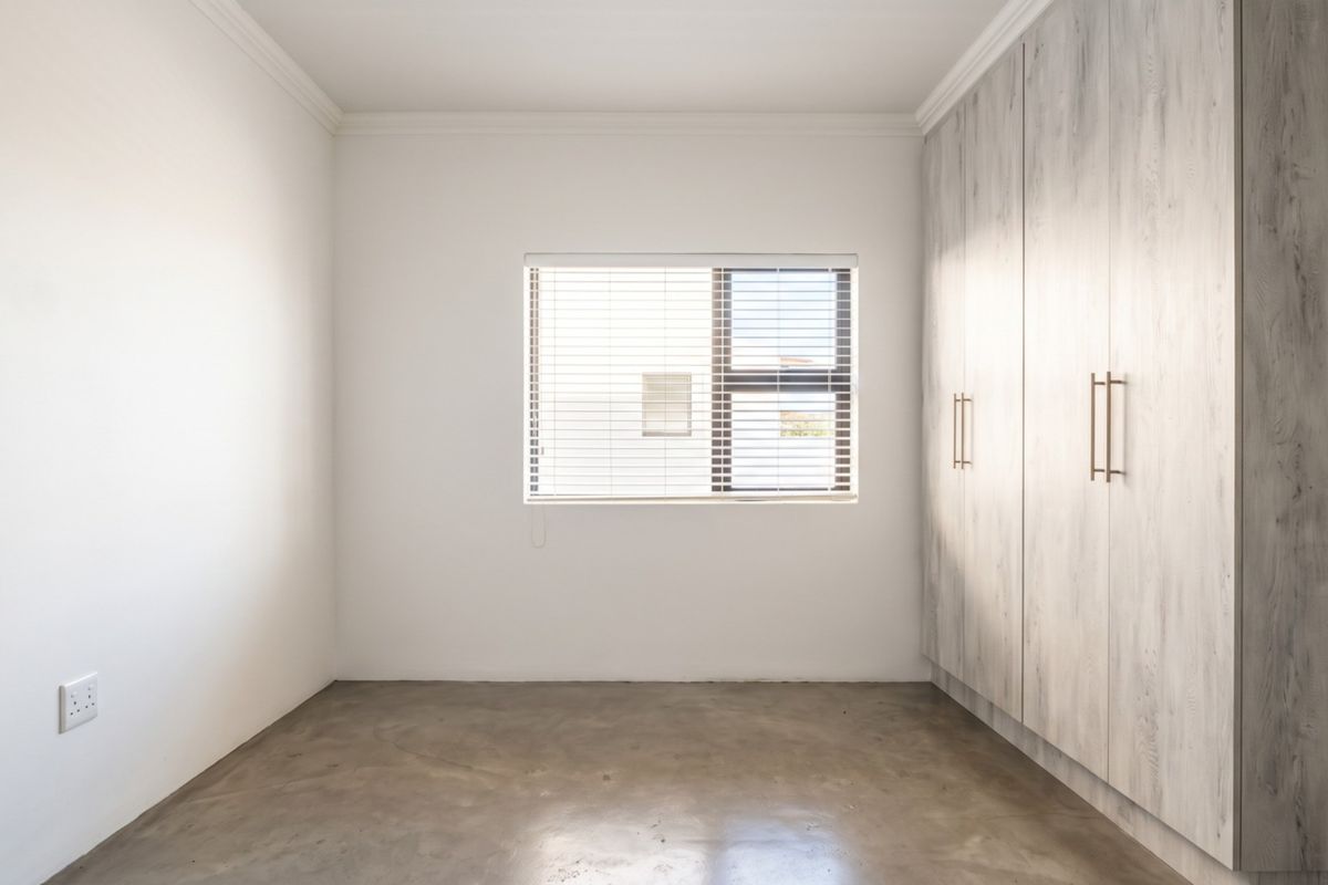 A bright, empty bedroom featuring polished concrete floors, built-in cupboards with a wood-grain finish, and a window fitted with venetian blinds.