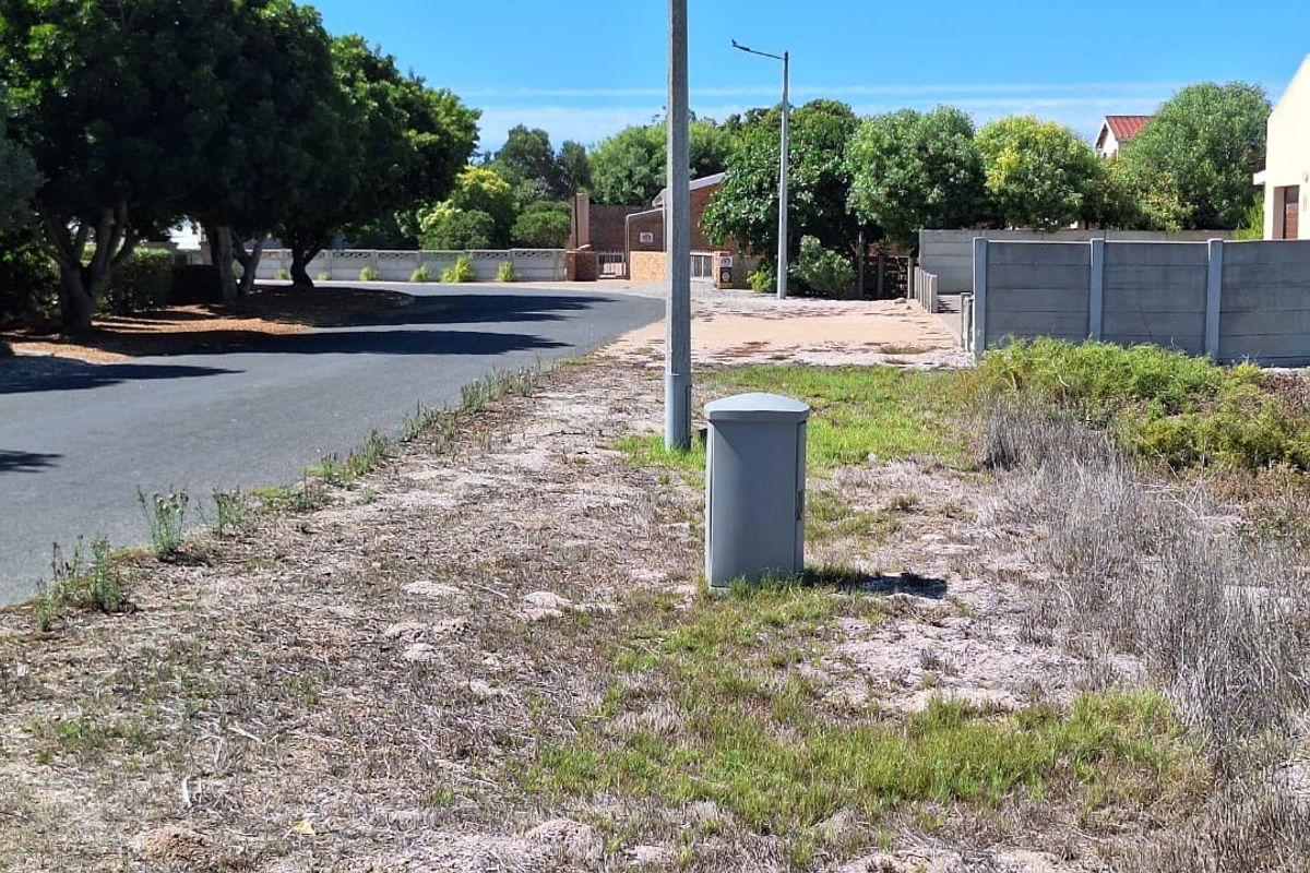A street-level view of the residential plot showing the adjacent asphalt road, boundary wall, and utility infrastructure.