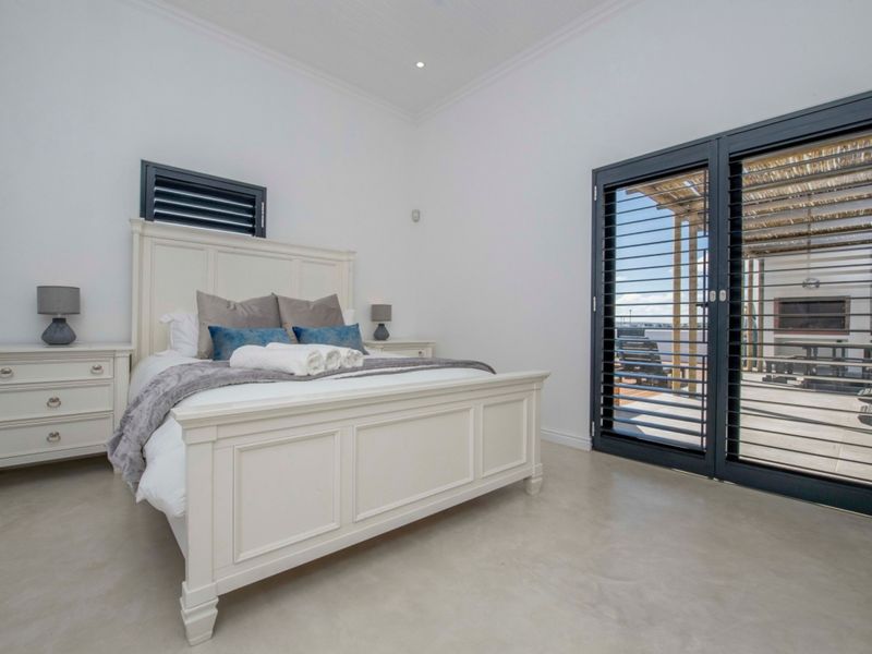 A bright, modern bedroom featuring polished concrete floors, recessed lighting, and large sliding doors with louvre shutters providing access to an outdoor patio area.