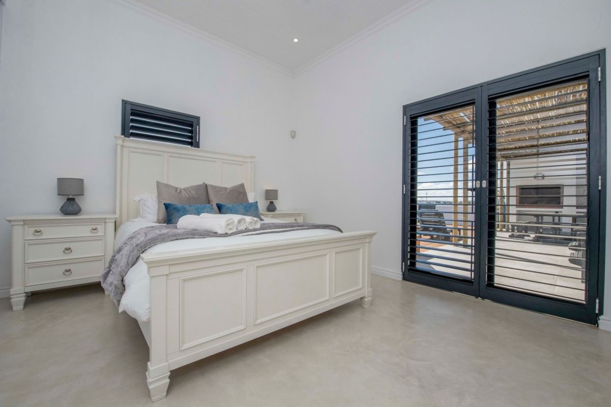 A bright, modern bedroom featuring polished concrete floors, recessed lighting, and large sliding doors with louvre shutters providing access to an outdoor patio area.