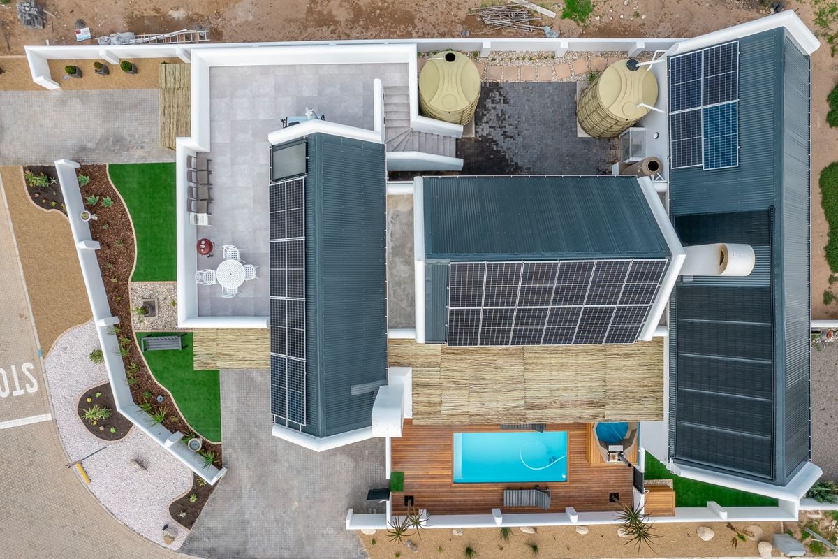 Aerial view of a modern property featuring extensive solar panel arrays, a swimming pool, timber decking, water storage tanks, and landscaped outdoor areas.