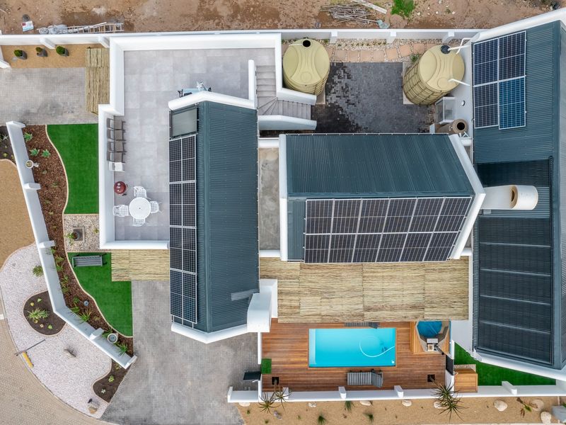 Aerial view of a modern property featuring extensive solar panel arrays, a swimming pool, timber decking, water storage tanks, and landscaped outdoor areas.