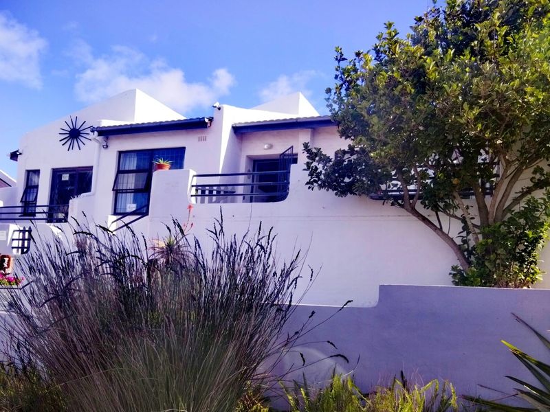 Front elevation of a white-painted house featuring metal balcony railings, casement windows, and a low boundary wall with established garden landscaping.