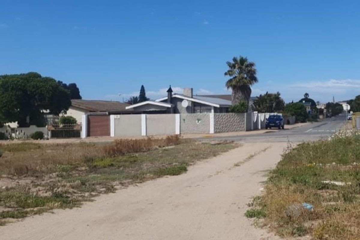A street-level view of a residential area showing a vacant dirt access path leading toward a paved road and neighboring properties with perimeter walls.