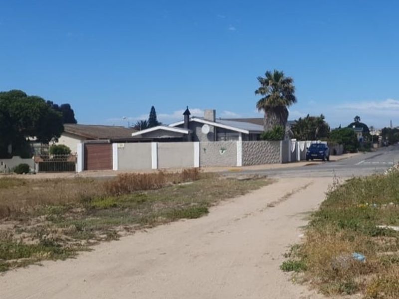 A street-level view of a residential area showing a vacant dirt access path leading toward a paved road and neighboring properties with perimeter walls.