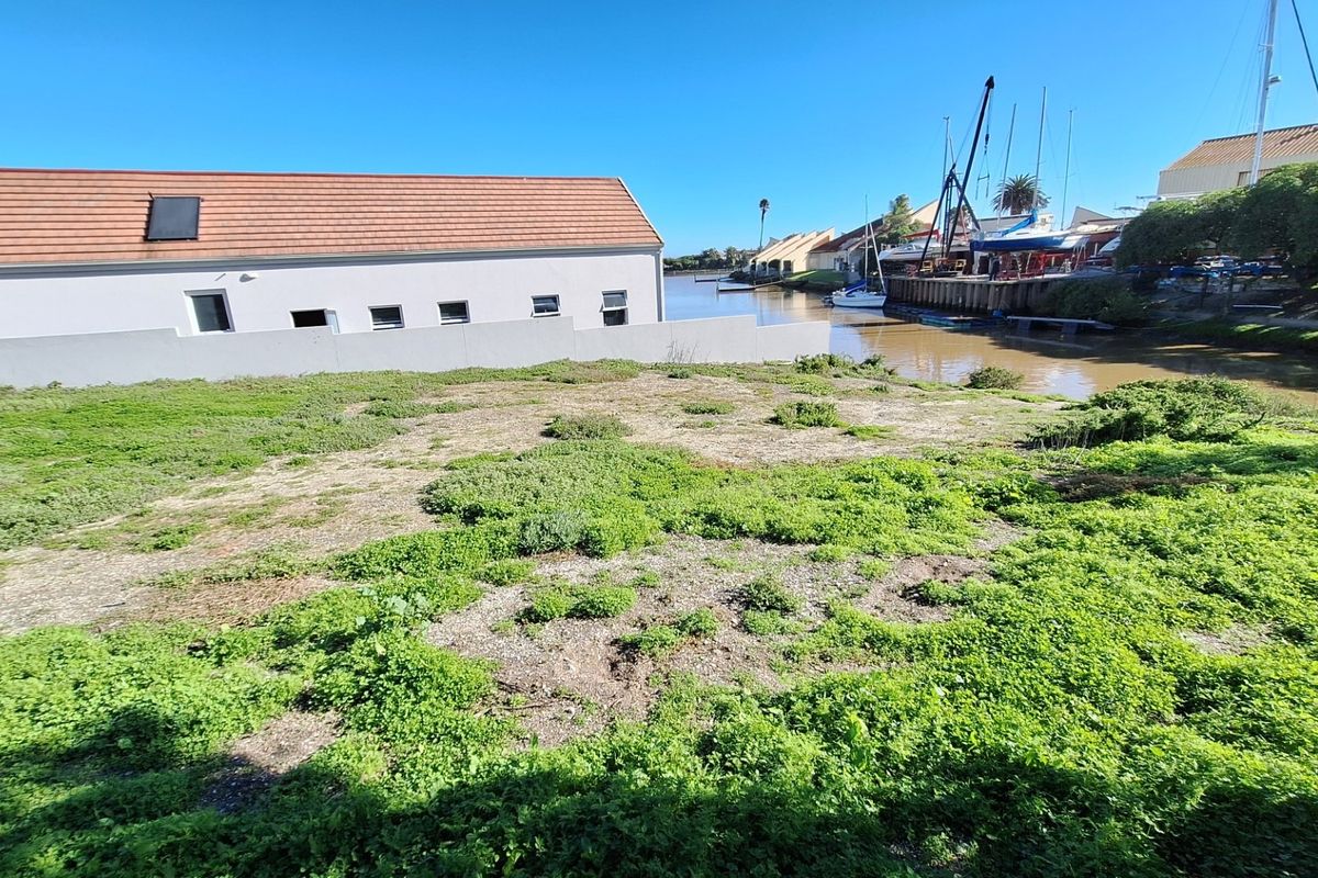 A vacant residential plot featuring direct waterfront access, situated adjacent to a canal with nearby boat moorings and established residential buildings.