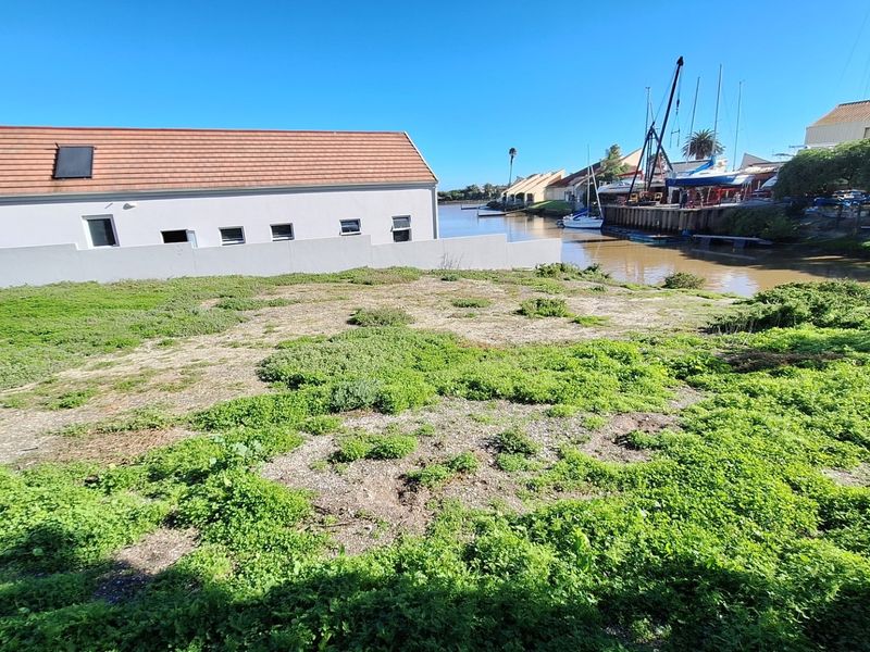 A vacant residential plot featuring direct waterfront access, situated adjacent to a canal with nearby boat moorings and established residential buildings.