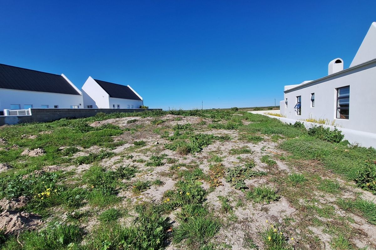 A vacant residential plot situated between existing modern white-walled homes, featuring natural ground cover and clear boundary lines.
