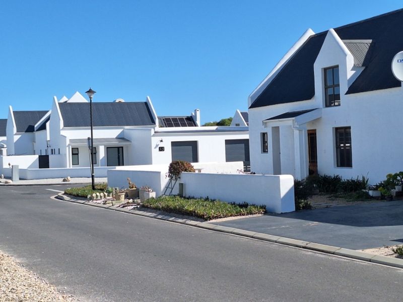 A street-level view of a residential estate featuring modern white homes with dark roofs, paved driveways, and well-maintained boundary walls.