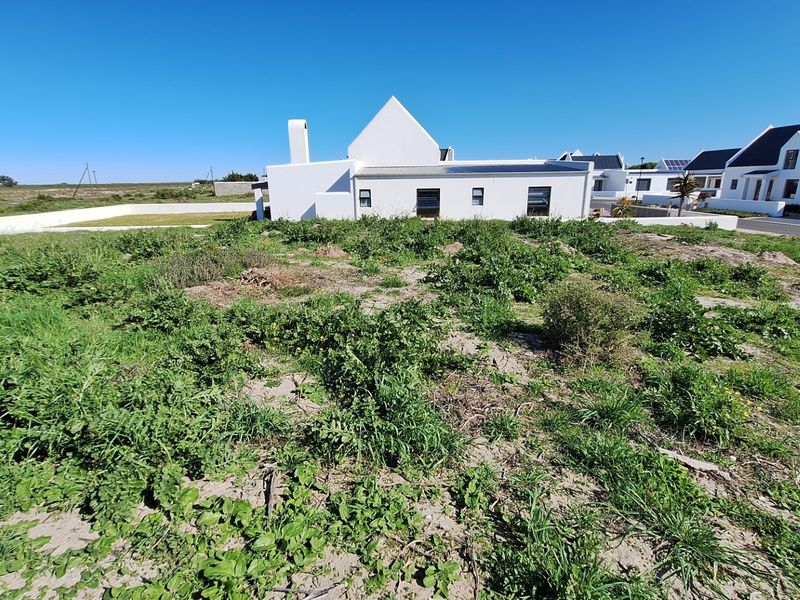 A vacant residential plot of land with natural vegetation, featuring a boundary wall and adjacent residential properties in a coastal estate setting.