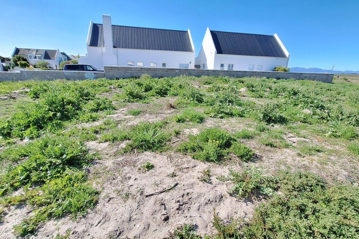 A vacant residential plot featuring sandy terrain and a boundary wall, with neighboring properties visible in the background.