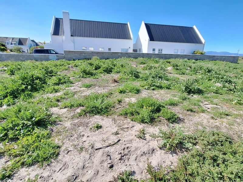 A vacant residential plot featuring sandy terrain and a boundary wall, with neighboring properties visible in the background.