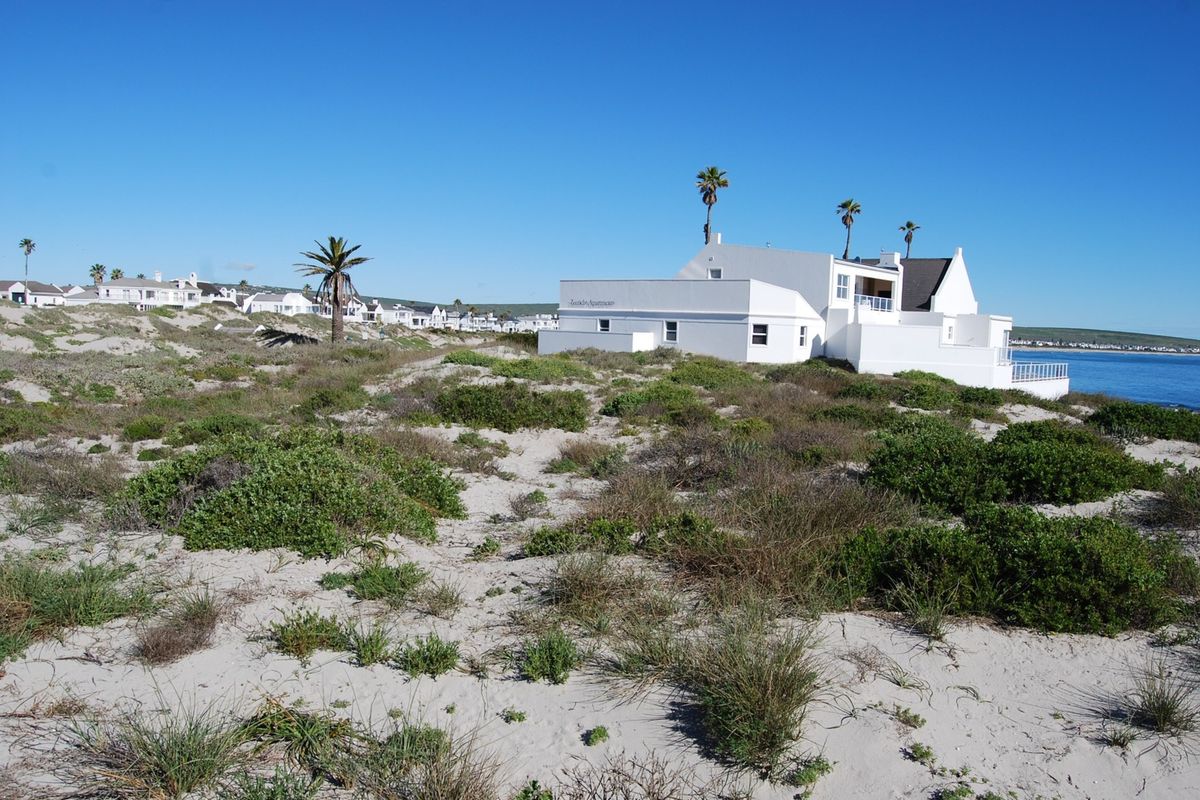 A view of a white coastal property situated on sandy dunes with native vegetation, overlooking the ocean.