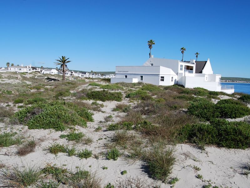 A view of a white coastal property situated on sandy dunes with native vegetation, overlooking the ocean.