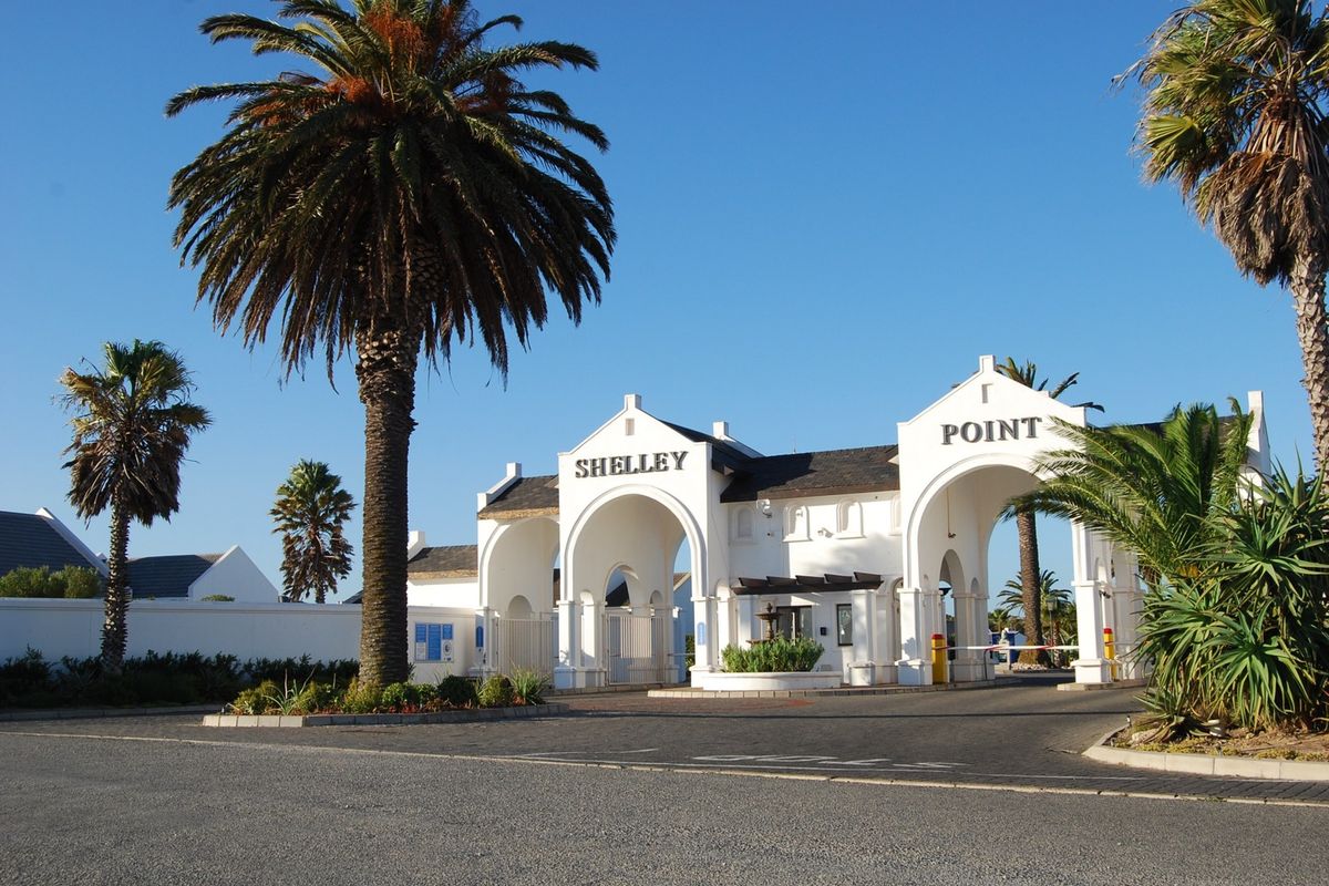 The main entrance gate to Shelley Point estate, featuring a white architectural archway, security boom, and well-maintained palm tree landscaping.