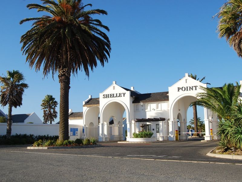 The main entrance gate to Shelley Point estate, featuring a white architectural archway, security boom, and well-maintained palm tree landscaping.