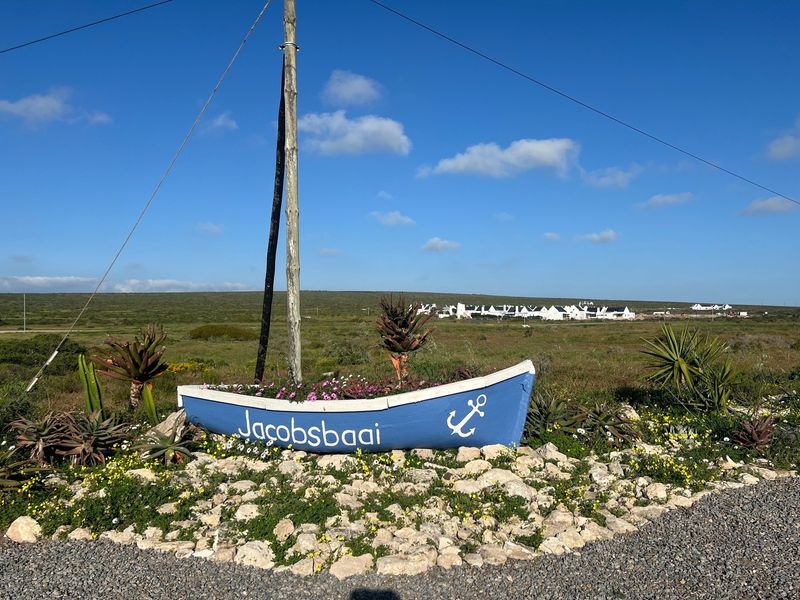 A decorative boat-shaped planter marking the entrance to Jacobsbaai, set against a backdrop of open residential land and coastal vegetation.