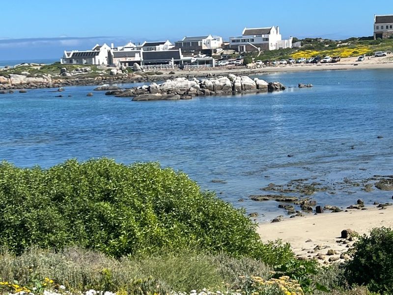 A scenic view of the coastal landscape featuring a rocky shoreline, sandy beach, and distant residential properties under a clear blue sky.