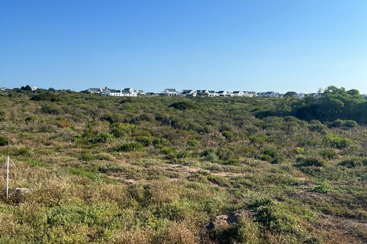 A view of a vacant residential plot featuring natural coastal vegetation and distant neighborhood housing under a clear blue sky.