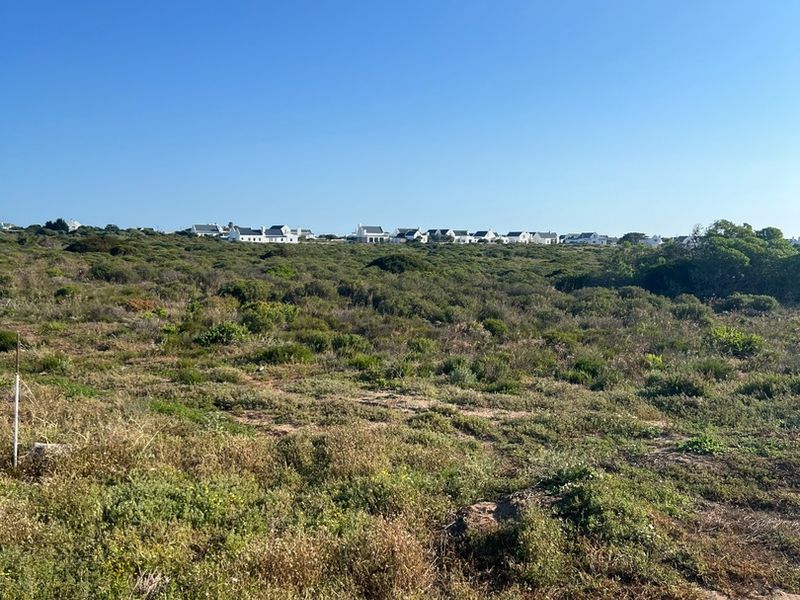 A view of a vacant residential plot featuring natural coastal vegetation and distant neighborhood housing under a clear blue sky.