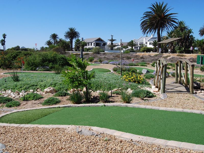 A well-maintained miniature golf course set within a landscaped garden, featuring a wooden footbridge and a central stone fountain.
