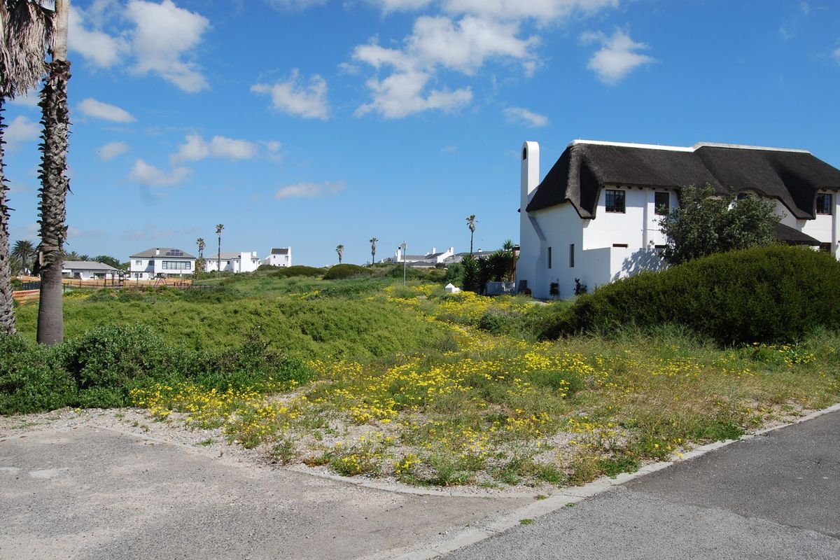 A view of a vacant residential plot featuring natural vegetation, with a neighboring thatched-roof property visible in the background.