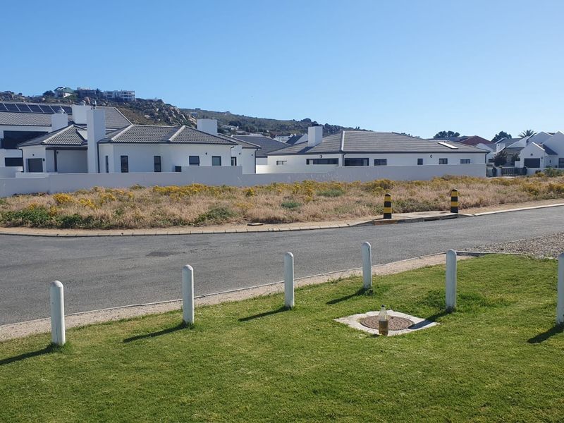 A view of a vacant residential plot featuring a paved road, concrete bollards, and a manicured grass verge in a developed neighborhood.